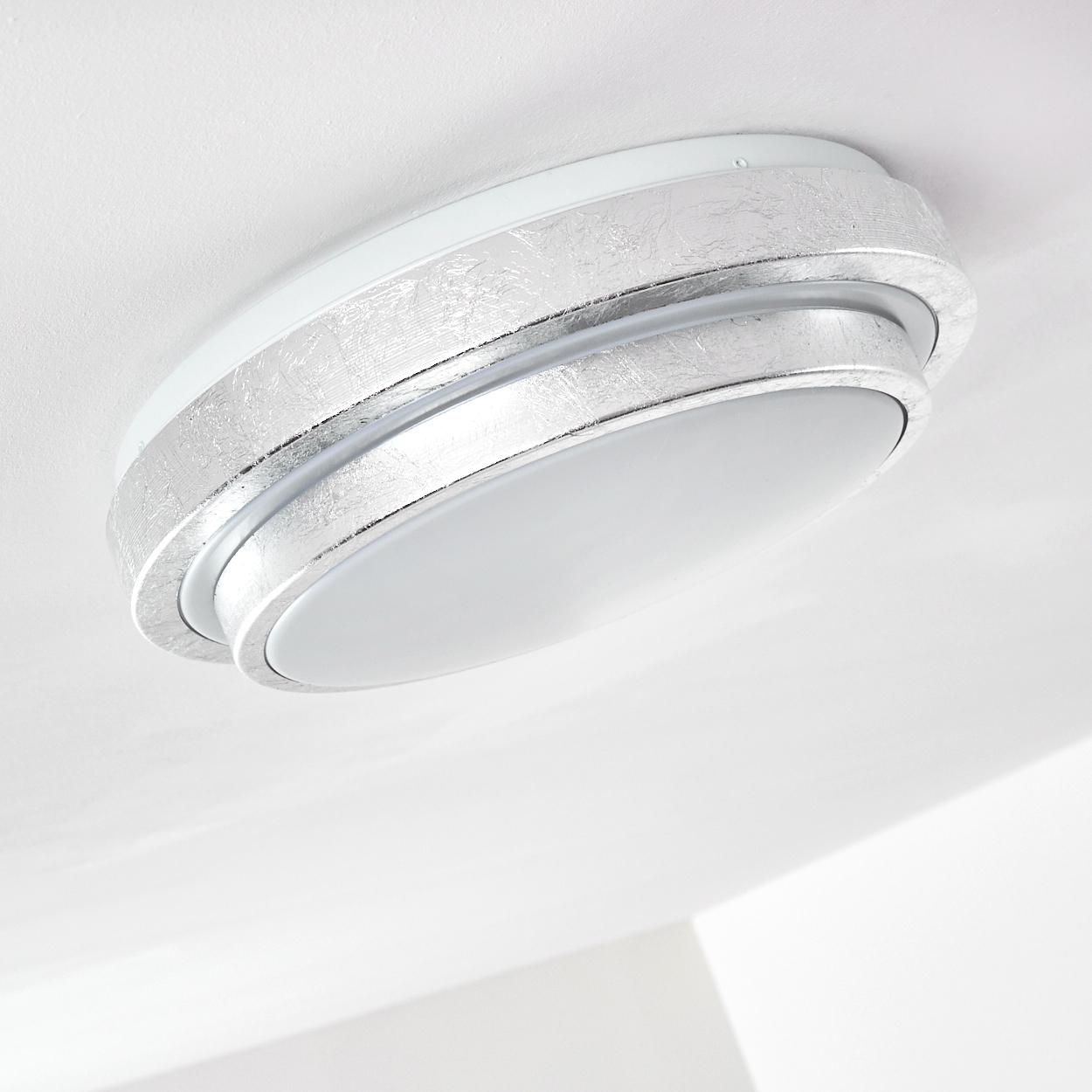 Eine moderne Deckenlampe mit silbernem Metallrand und weißer Lichtabdeckung, vor weißer Decke.