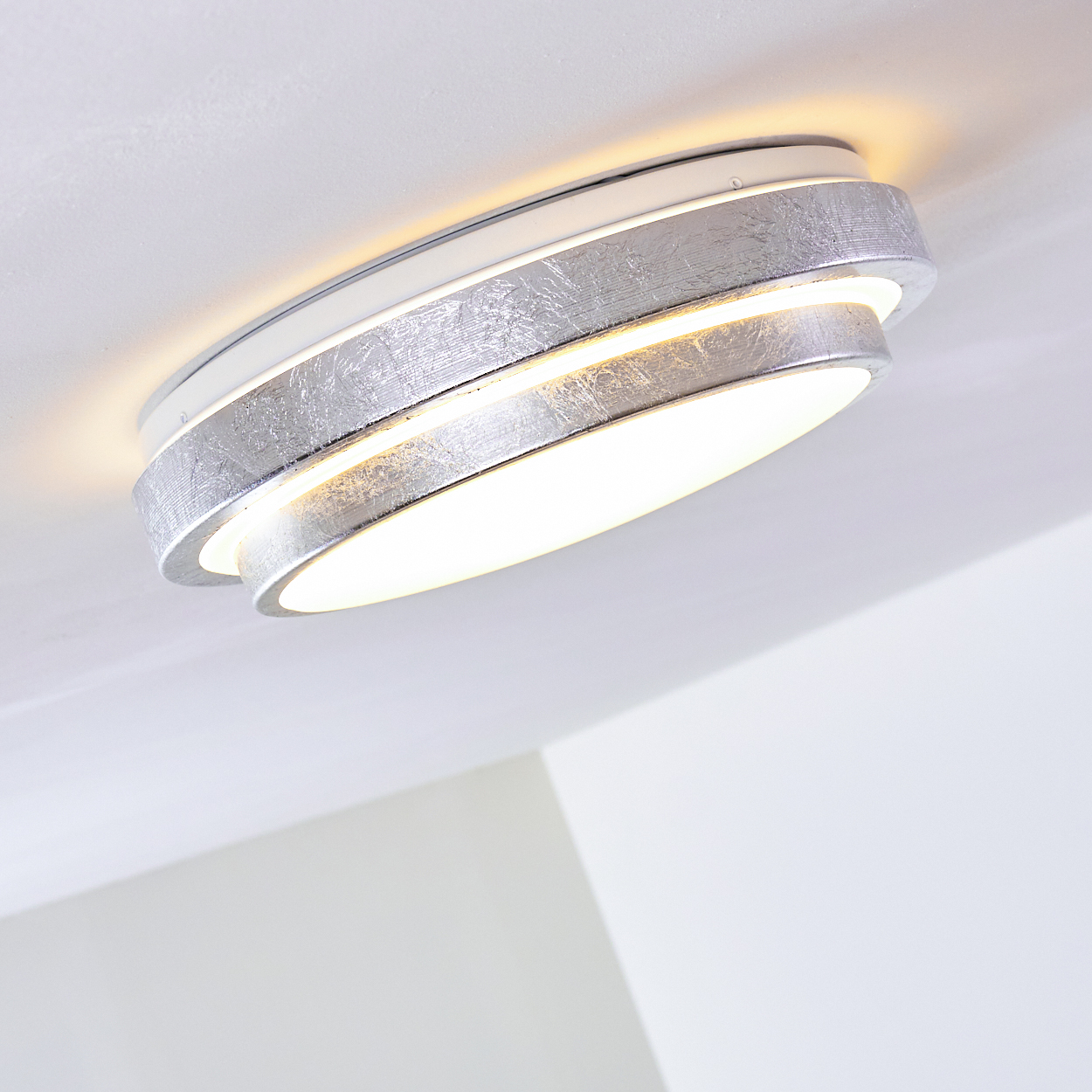 Eine moderne, silberne Deckenleuchte ist angewinkelt. Sie hat eine strukturierte Oberfläche und strahlt ein warmes Licht aus. Der Hintergrund ist eine weiße Decke.