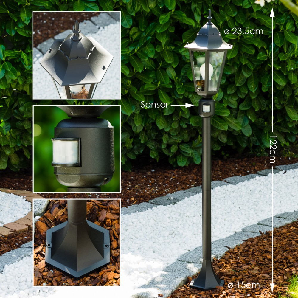 Eine schwarze Außenlampe mit Sensor. Sie steht auf einem Kiesweg, im Hintergrund ein grüner Busch. Abmessungen sind angegeben.