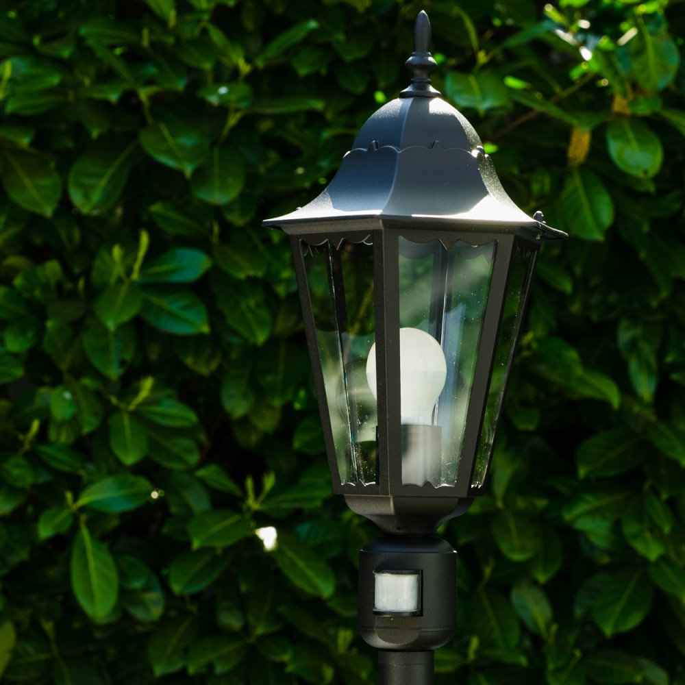 Schwarze Außenlampe mit einer weißen Glühbirne, vor einem grünen, belaubten Hintergrund. Sie hat einen Bewegungssensor.