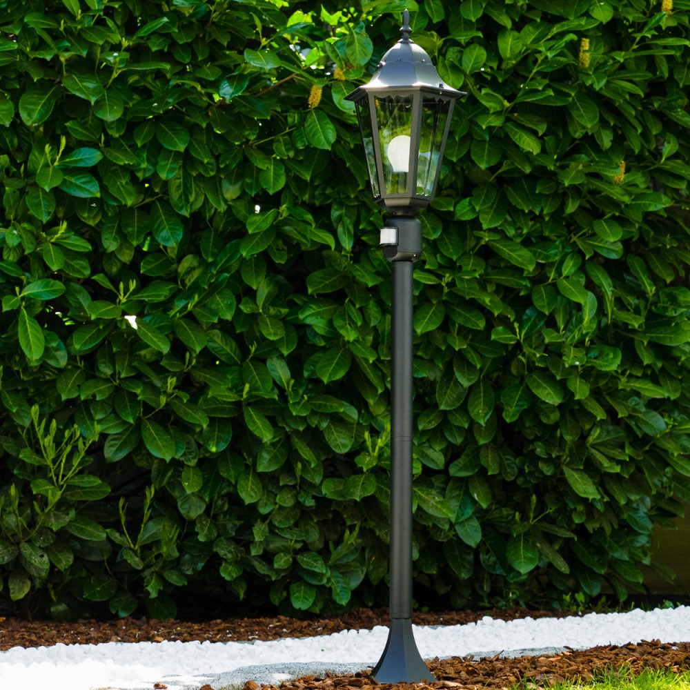 Eine schwarze Laterne steht vor einer dichten grünen Hecke. Weiße Steine säumen die Basis der Hecke. Die Lampe hat eine Glühbirne.