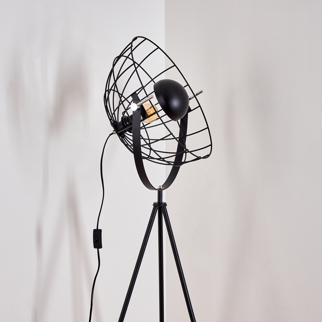 Eine schwarze Stehlampe mit käfigartigem Lampenschirm, dreibeinigem Sockel und Netzkabel vor einer weißen Wand in der Ecke.