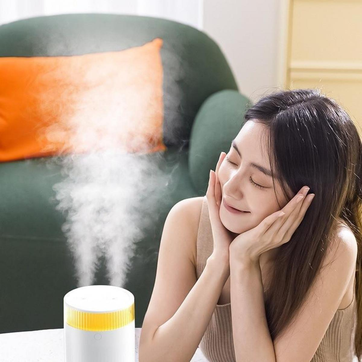 Eine Frau mit geschlossenen Augen und Händen auf den Wangen genießt einen Luftbefeuchter, der Nebel ausstößt. Grünes Sofa und orangefarbenes Kissen im Hintergrund.