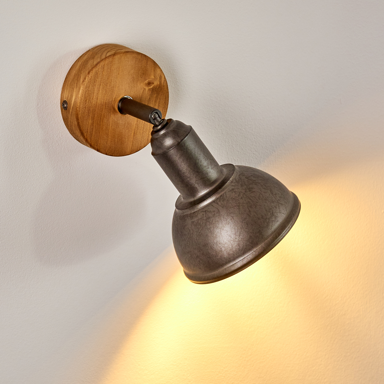 Eine Wandleuchte aus Metall und Holz. Die Lampe ist nach unten gerichtet und beleuchtet die weiße Wand. Der Holzteil ist rund.