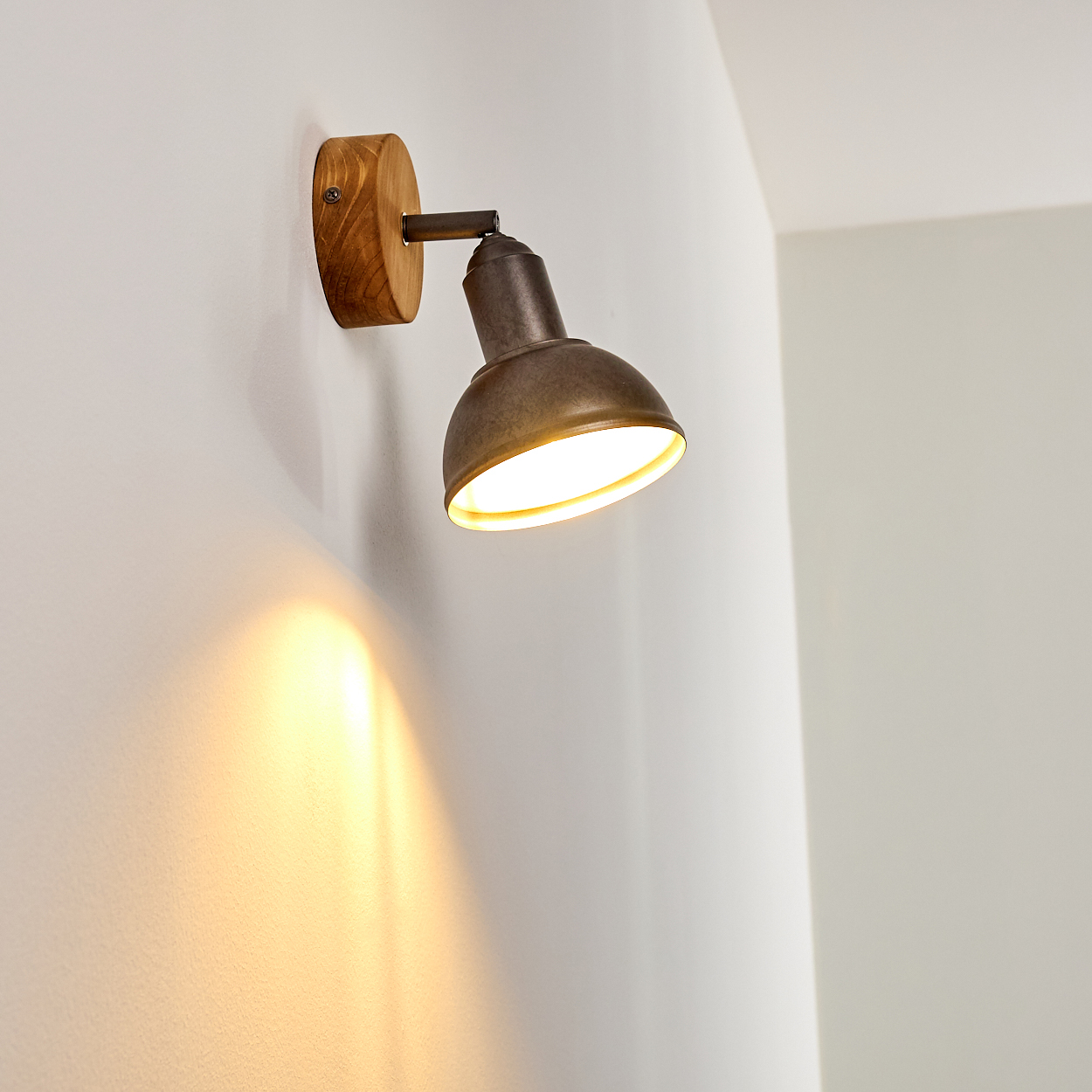 Eine Wandlampe mit Holzsockel und metallischem Lampenschirm beleuchtet eine weiße Wand. Licht reflektiert von der Wand.