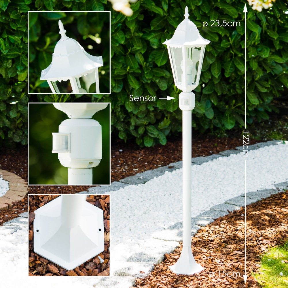Eine weiße Außenlaterne mit Sensor. Sie steht in einem Garten mit Pflanzen und weißen Steinen. Enthält Details der Lampenteile.