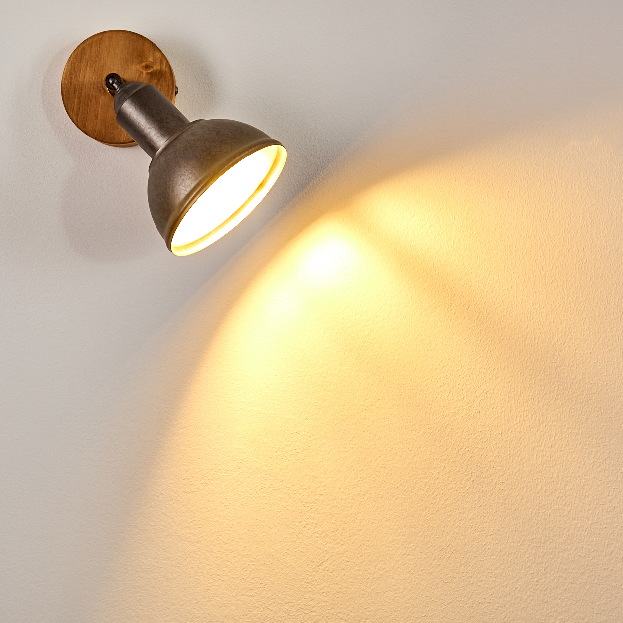 Ein Metall-Spotlicht auf Holzgrund leuchtet auf eine strukturierte weiße Wand. Das Licht erzeugt einen warmen, gelben Schein.