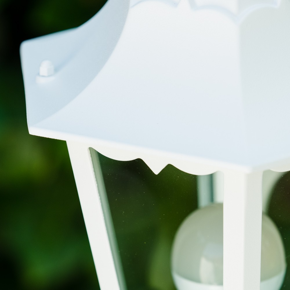 Nahaufnahme einer weißen Außenlampe. Die Lampe hat ein dekoratives Dach und Glasscheiben mit einer Glühbirne im Inneren. Verschwommener grüner Hintergrund.