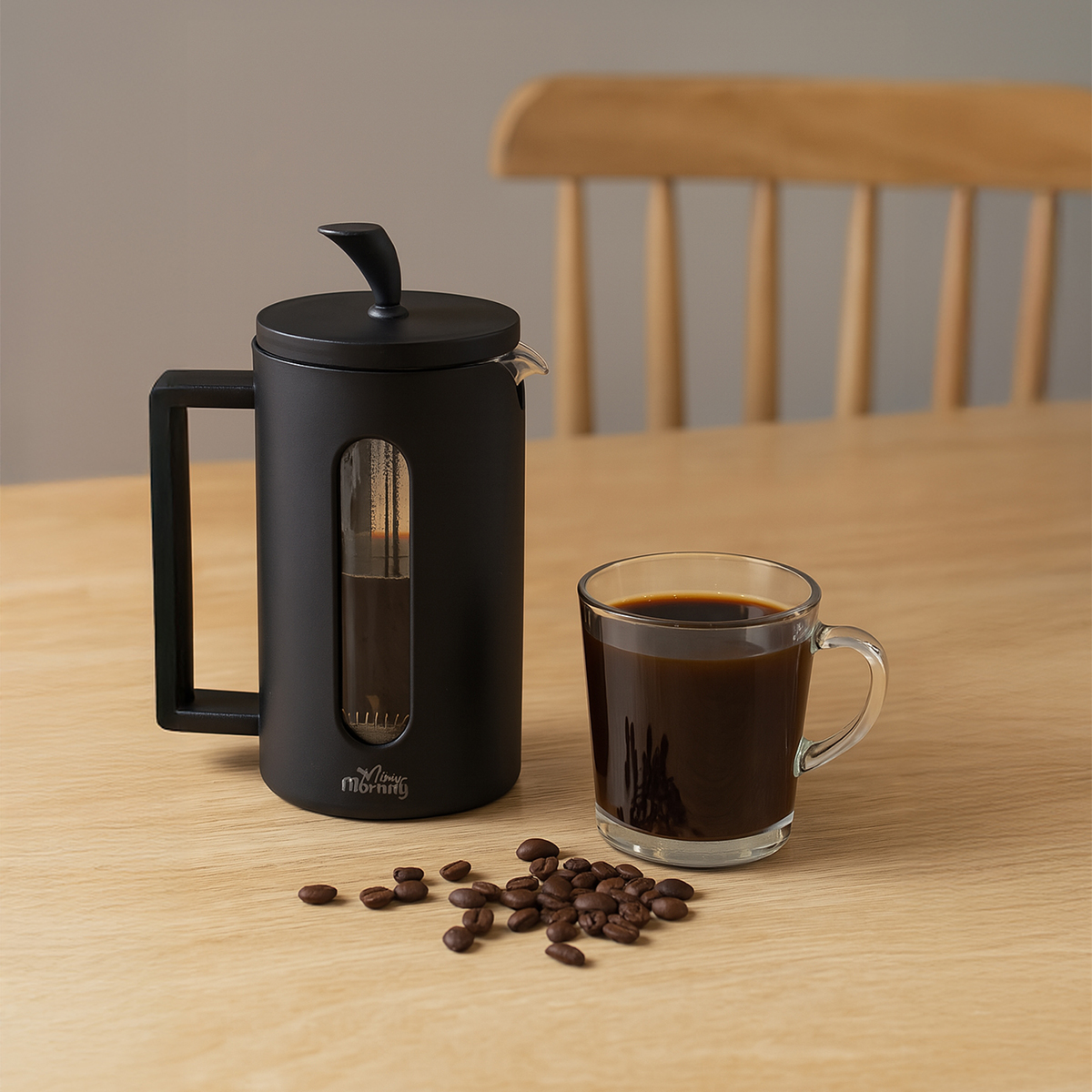 Una prensa francesa y una taza de café en una mesa de madera con granos de café. Una silla en el fondo borroso.