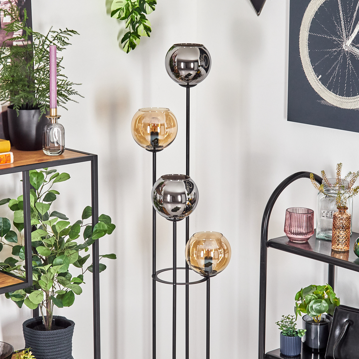 Eine dekorative Lampe mit vier Glaskugeln. Regale mit Pflanzen, Kerzen und Dekorationsgegenständen. Fahrradradkunst an der Wand.