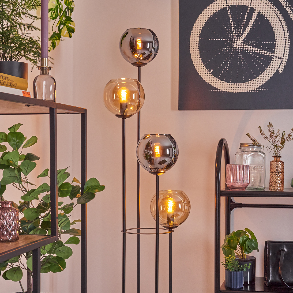 Eine Stehlampe mit vier Glaskugeln leuchtet. Regale halten Pflanzen und Dekorationsartikel. Ein Fahrradrad-Kunstwerk hängt an der Wand.