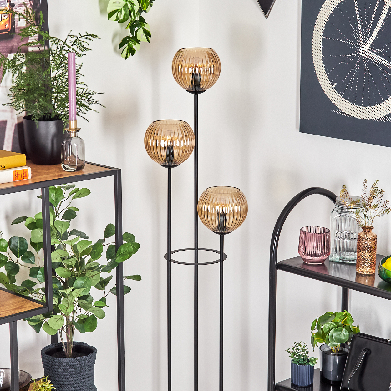 Ein Raum mit Regalen, Pflanzen und einer dreiflammigen Lampe. Regale halten Bücher, Kerzen und Vasen. An der Wand befindet sich ein Fahrradrad-Kunstwerk.