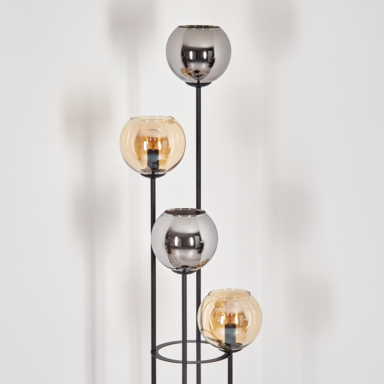 Eine moderne Lampe mit vier Glaskugeln, abwechselnd silber und bernsteinfarben, auf einem schwarzen Metallrahmen, vor einer weißen Wand.
