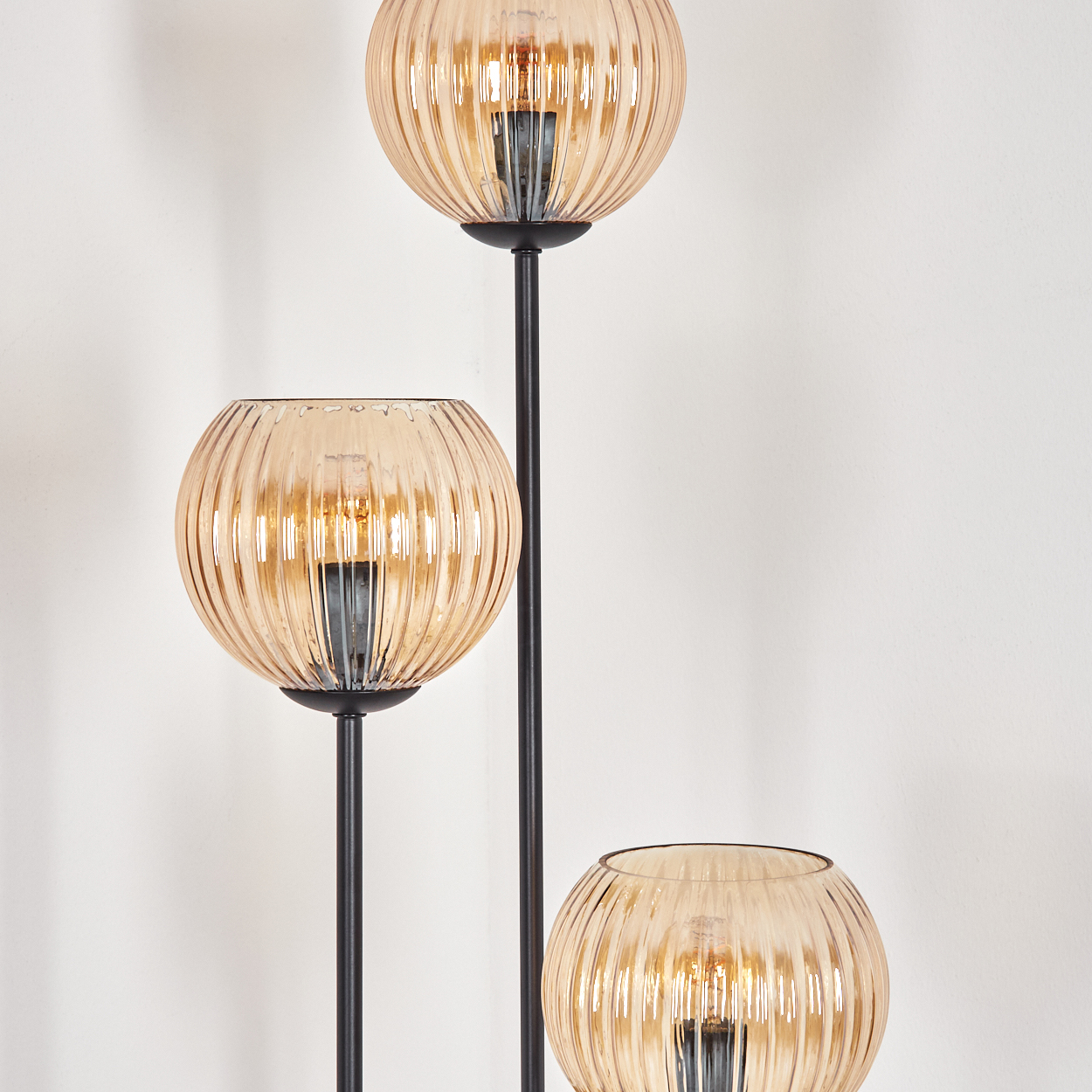 Stehlampe mit drei bernsteinfarbenen, gerippten Glaskugeln. Schwarzer Metallrahmen. Weißer Wandhintergrund.