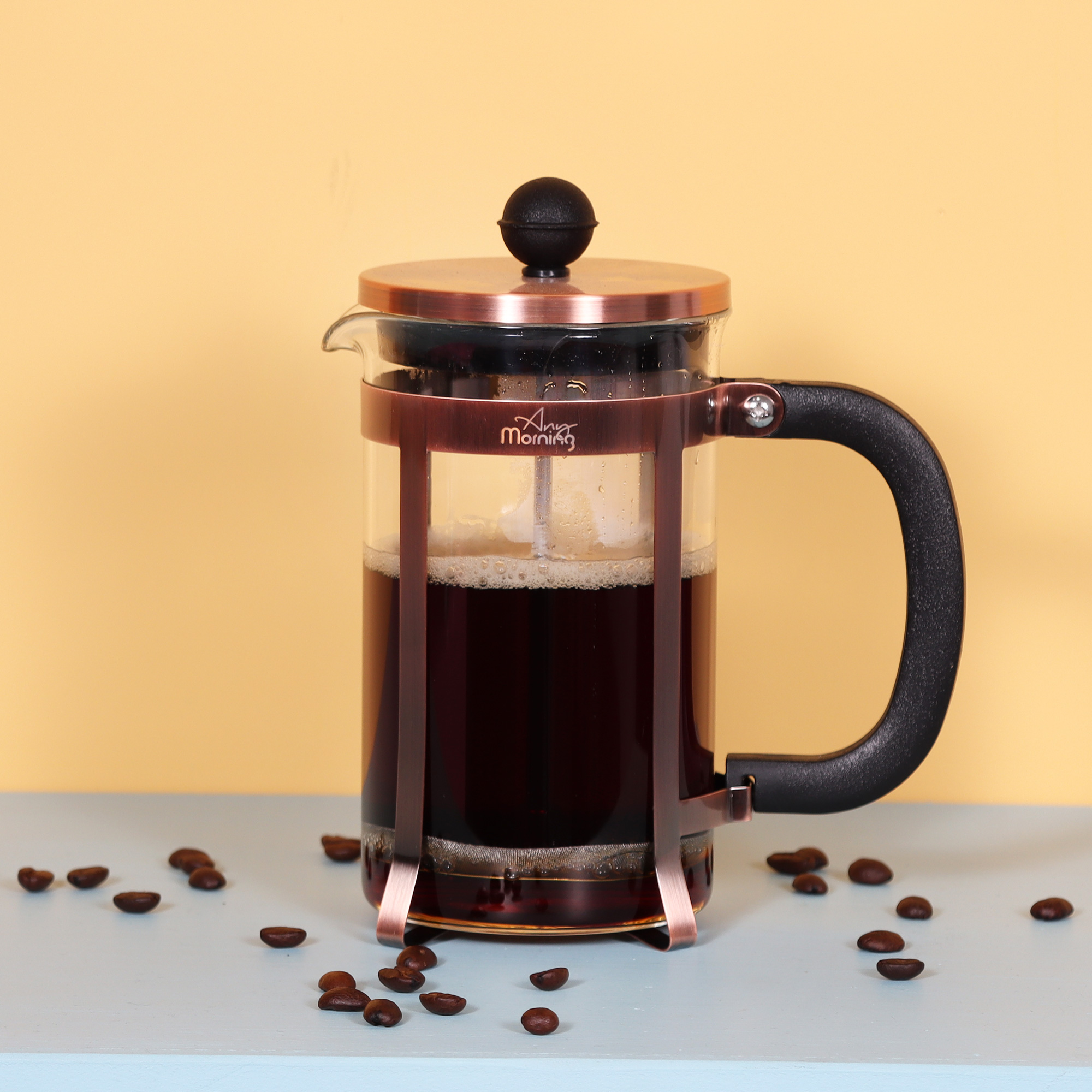 Una prensa francesa llena de café, detalles de cobre, mango y tapa negros. Granos de café esparcidos.