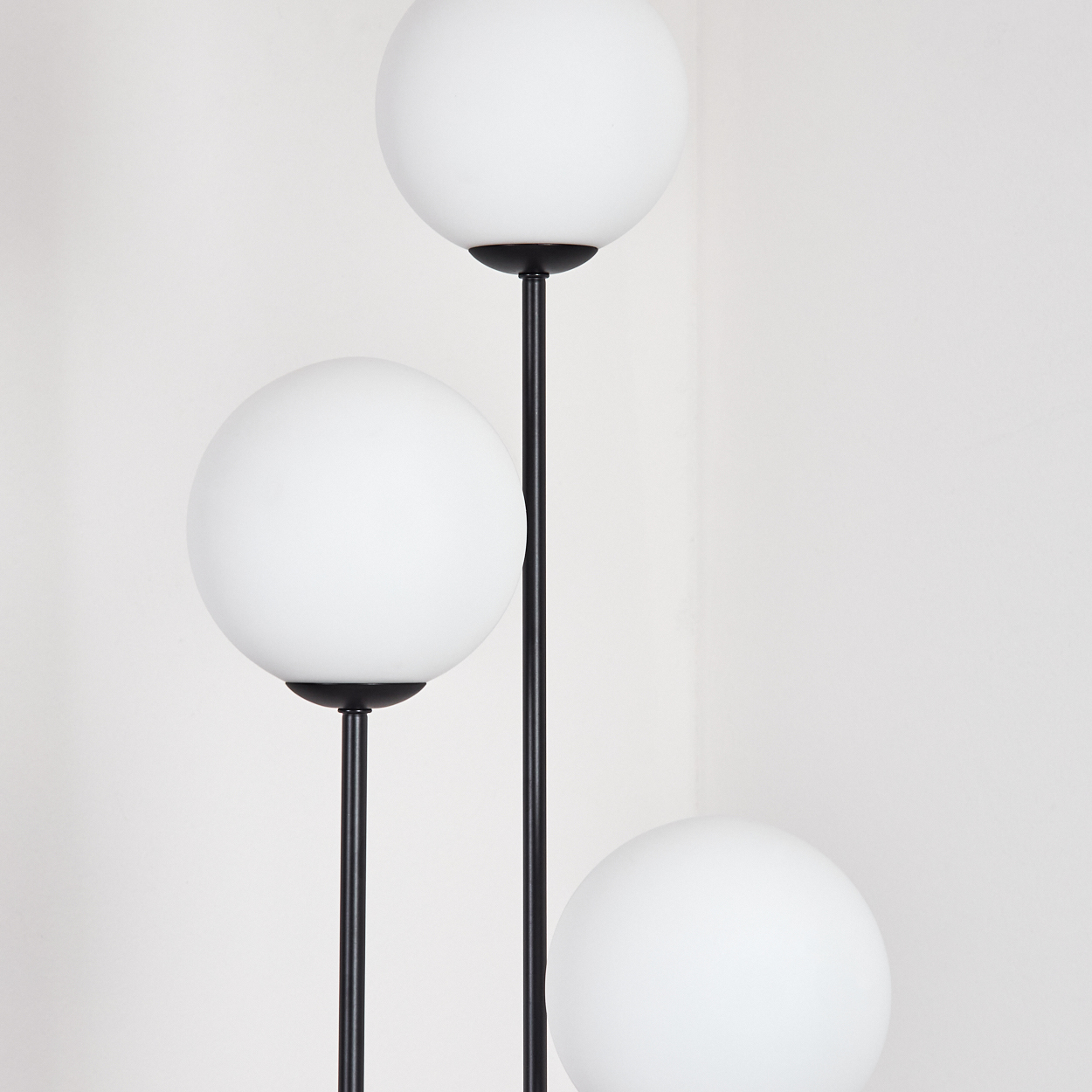 Eine moderne Stehlampe mit drei kugelförmigen weißen Glasschirmen und schwarzen Stielen vor einer weißen Wand.