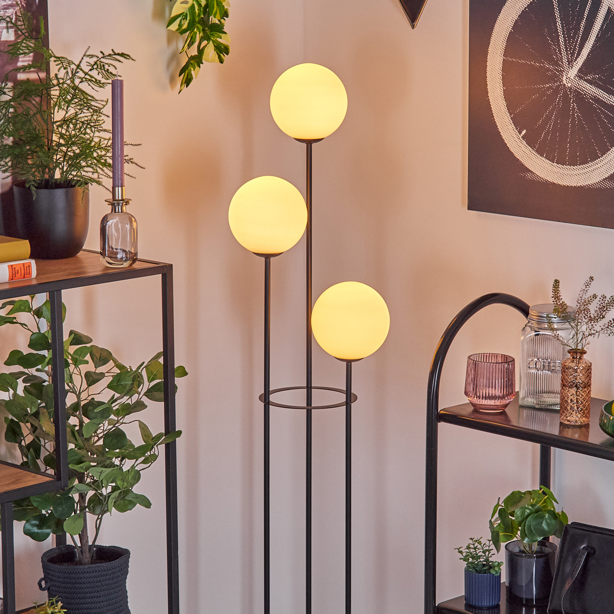 Eine moderne Zimmerecke mit einer dreiflammigen Stehlampe, Pflanzen, Regalen und Dekorationsartikeln. Die Wände sind hellrosa.