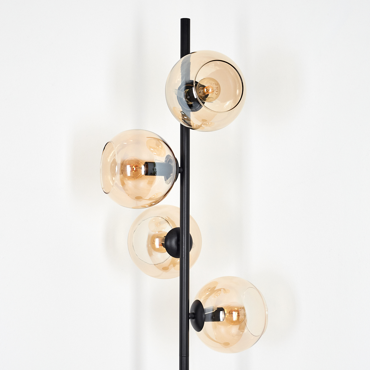 Eine moderne Stehlampe mit vier bernsteinfarbenen Glaskugeln. Der schwarze Metallrahmen hält die Kugeln vor einer weißen Wand.