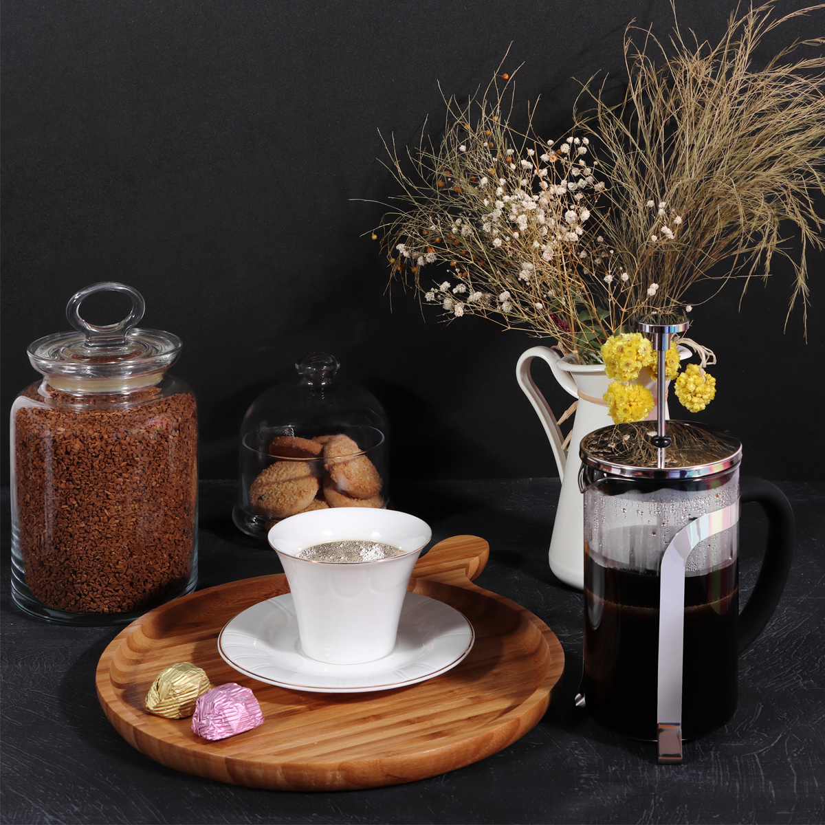 Preparación de café con taza, galletas, flores y prensa francesa en una bandeja de madera. También hay bombones.