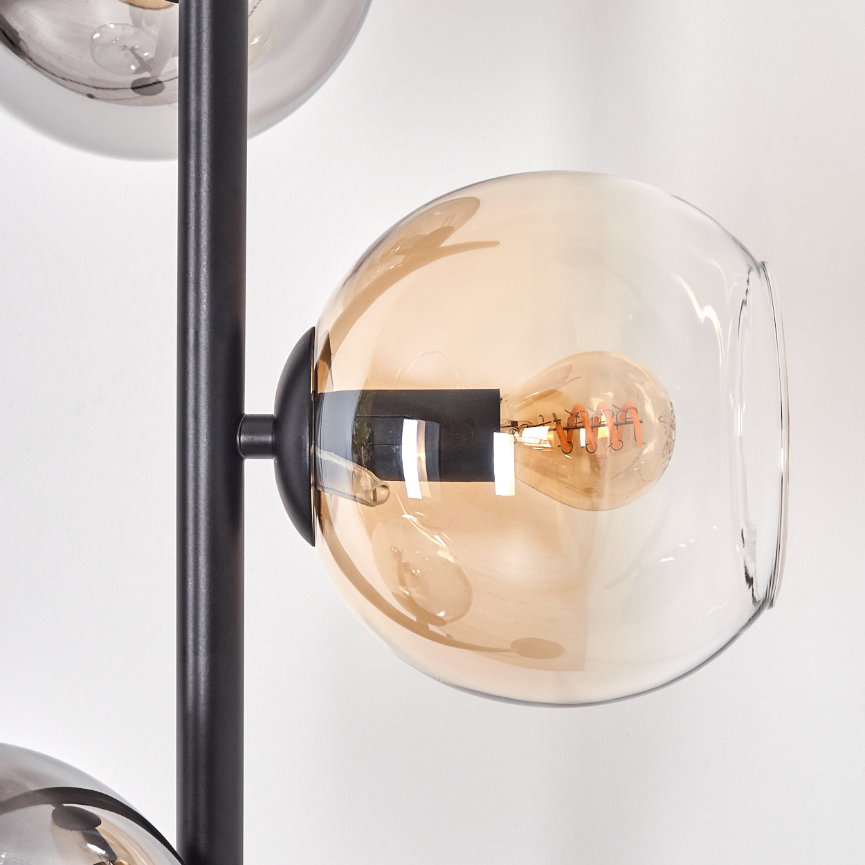 Eine moderne Lampe mit schwarzem Rahmen und Glaskugeln. Eine bernsteinfarbene Kugel beherbergt eine Glühbirne. Hintergrund ist eine weiße Wand.
