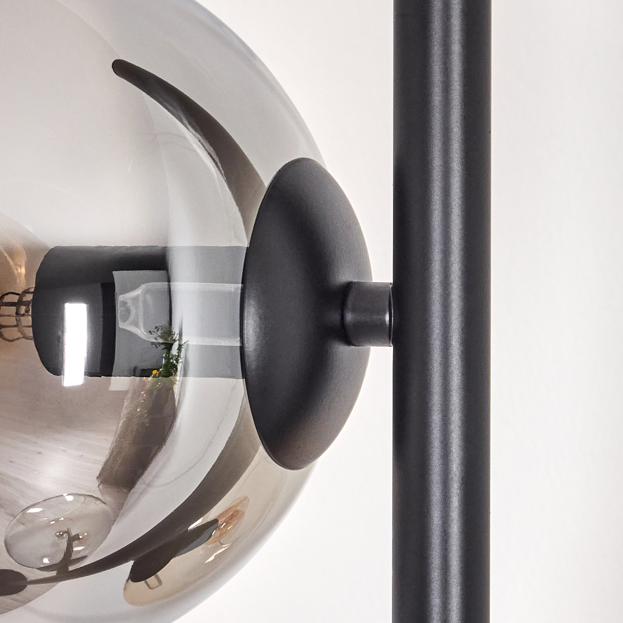 Eine moderne Lampe mit einer rauchfarbenen Glaskugel und einem schwarzen Metallrahmen. Reflexionen zeigen eine Rauminnenausstattung. Weißer Wandhintergrund.