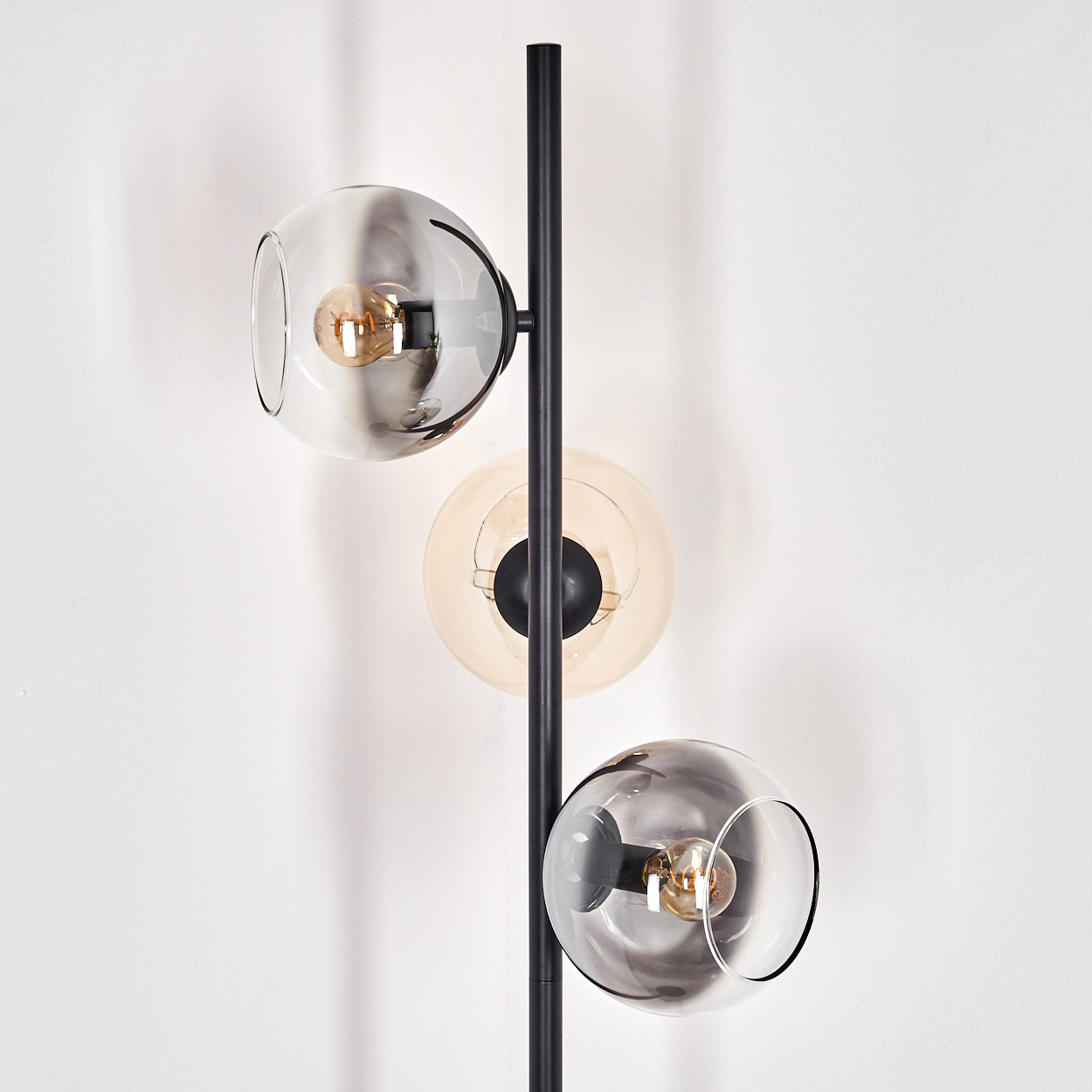 Eine moderne Stehlampe mit drei Glaskugelschirmen in Grau und Bernstein vor einer weißen Wand. Die Lampe hat eine schwarze Stange.