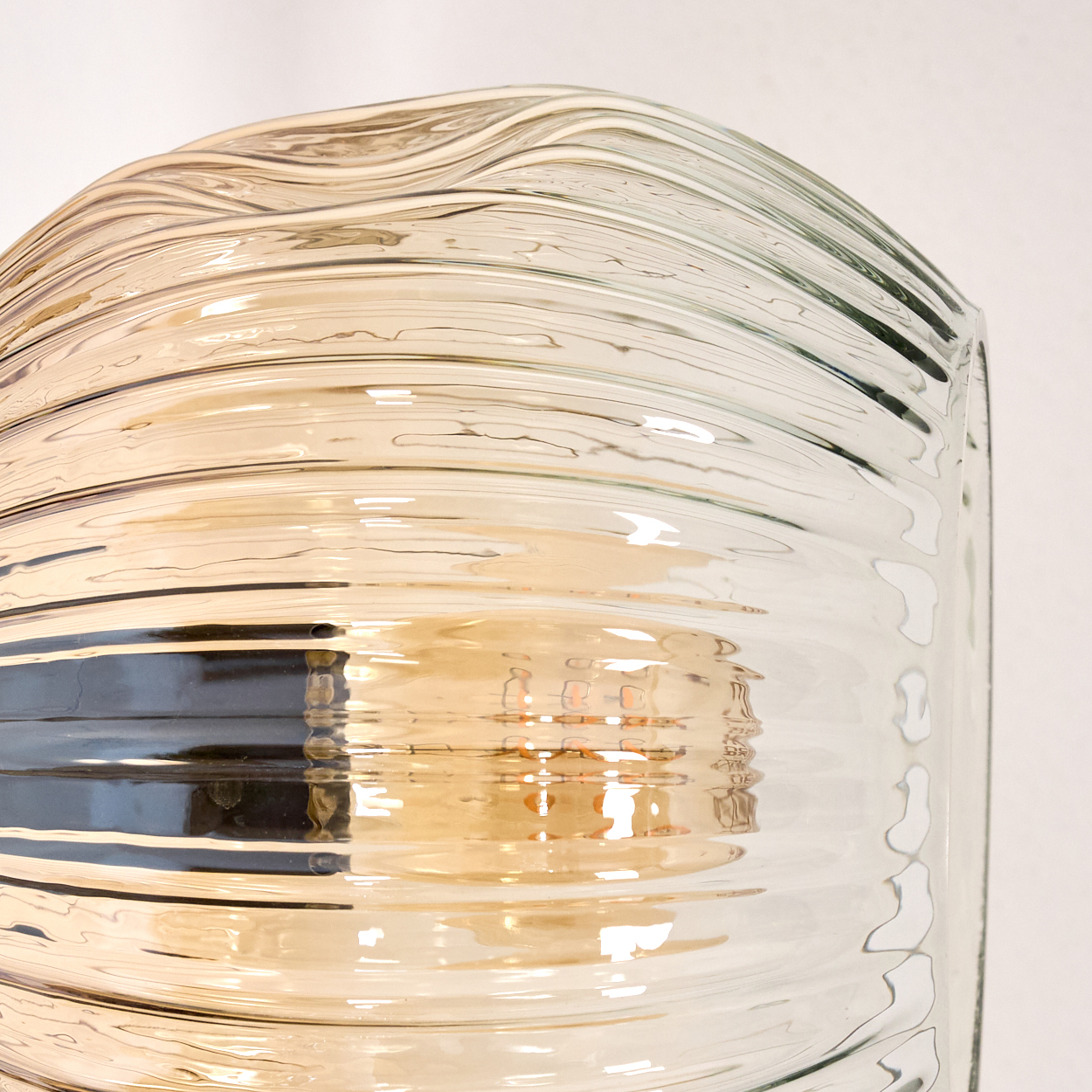Nahaufnahme eines Lampenschirms aus Glas mit horizontalen Rippen. Er ist klar mit einem gelblichen Farbton und reflektiert warmes Licht.