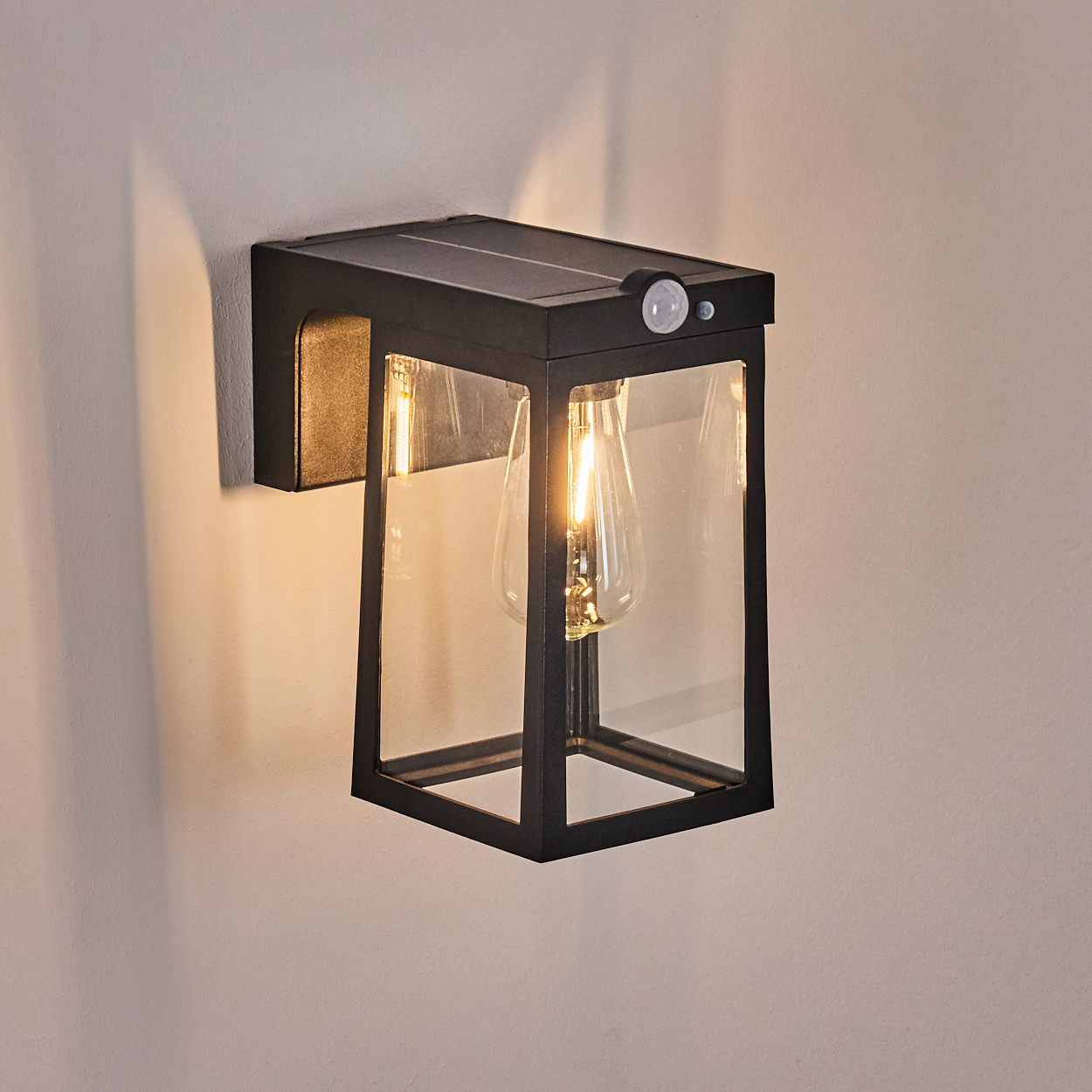 Eine schwarze Außenwandleuchte mit Solarpanel. Sie hat ein Glasgehäuse mit sichtbarer Glühbirne.