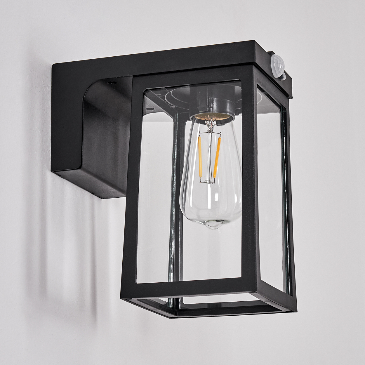 Schwarze Außenwandleuchte mit Glasgehäuse. Im Inneren befindet sich eine Glühbirne. Die Lampe ist an einer weißen Wand montiert.