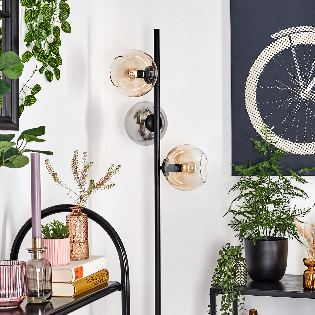 Eine moderne Stehlampe mit drei Glaskugeln. Verschiedene Pflanzen und Dekorationsartikel sind auf Regalen und Wänden angeordnet. Ein Fahrradkunstwerk.