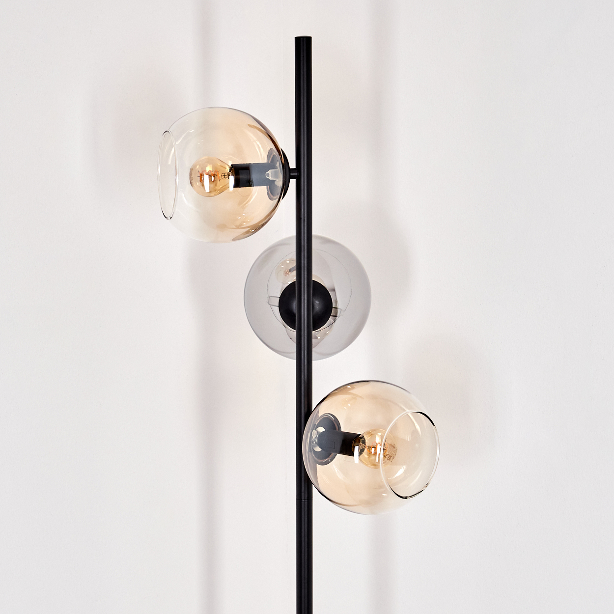 Eine moderne Stehlampe mit drei Glaskugel-Schirmen. Die Schirme sind bernsteinfarben und klar, mit schwarzem Metallträger. Der Hintergrund ist eine weiße Wand.