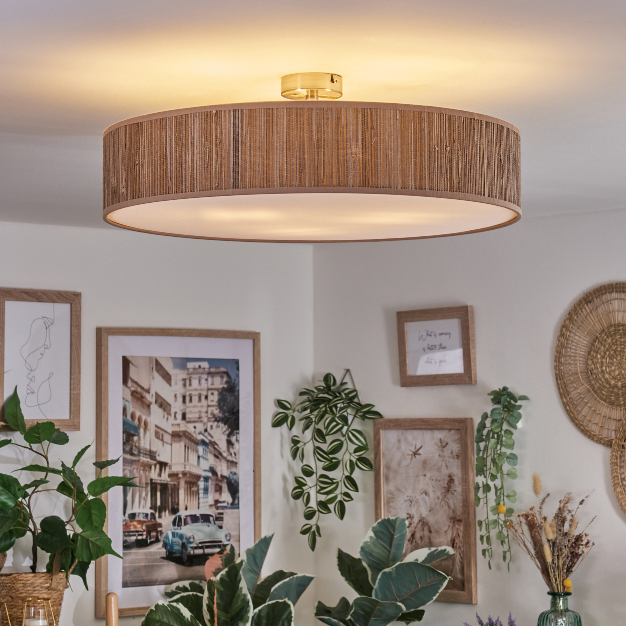 Eine braune, runde Deckenlampe hängt über gerahmten Bildern und Pflanzen. Die Bilder zeigen Stadtlandschaften und abstrakte Kunst. Wanddekoration.