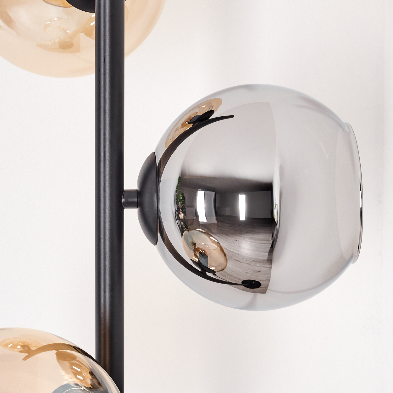 Moderne Lampe mit schwarzem Rahmen und drei kugelförmigen Glasschirmen, mit Spiegel- und Bernsteintönen, vor weißer Wand.