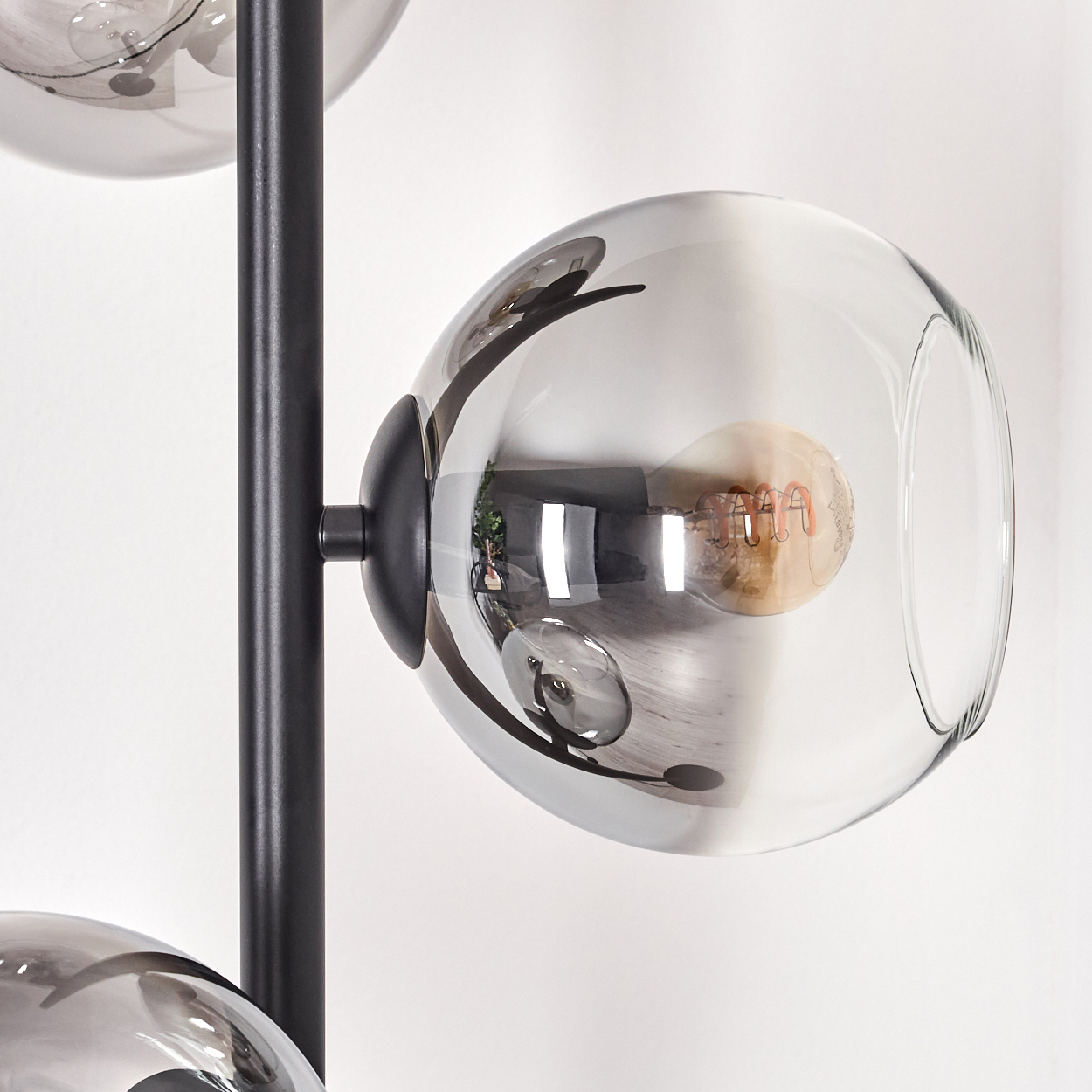 Eine moderne Lampe mit drei Glaskugeln. Jede Kugel enthält eine Glühbirne. Die Lampe hat ein schwarzes Metallgestell, vor einer weißen Wand.