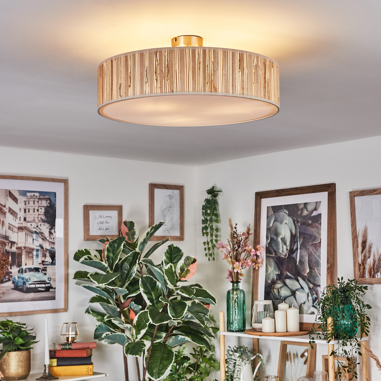 Ein Raum mit einer großen Deckenleuchte, gerahmten Bildern, grünen Pflanzen und einem kleinen Regal mit Kerzen und einer Vase mit Blumen.