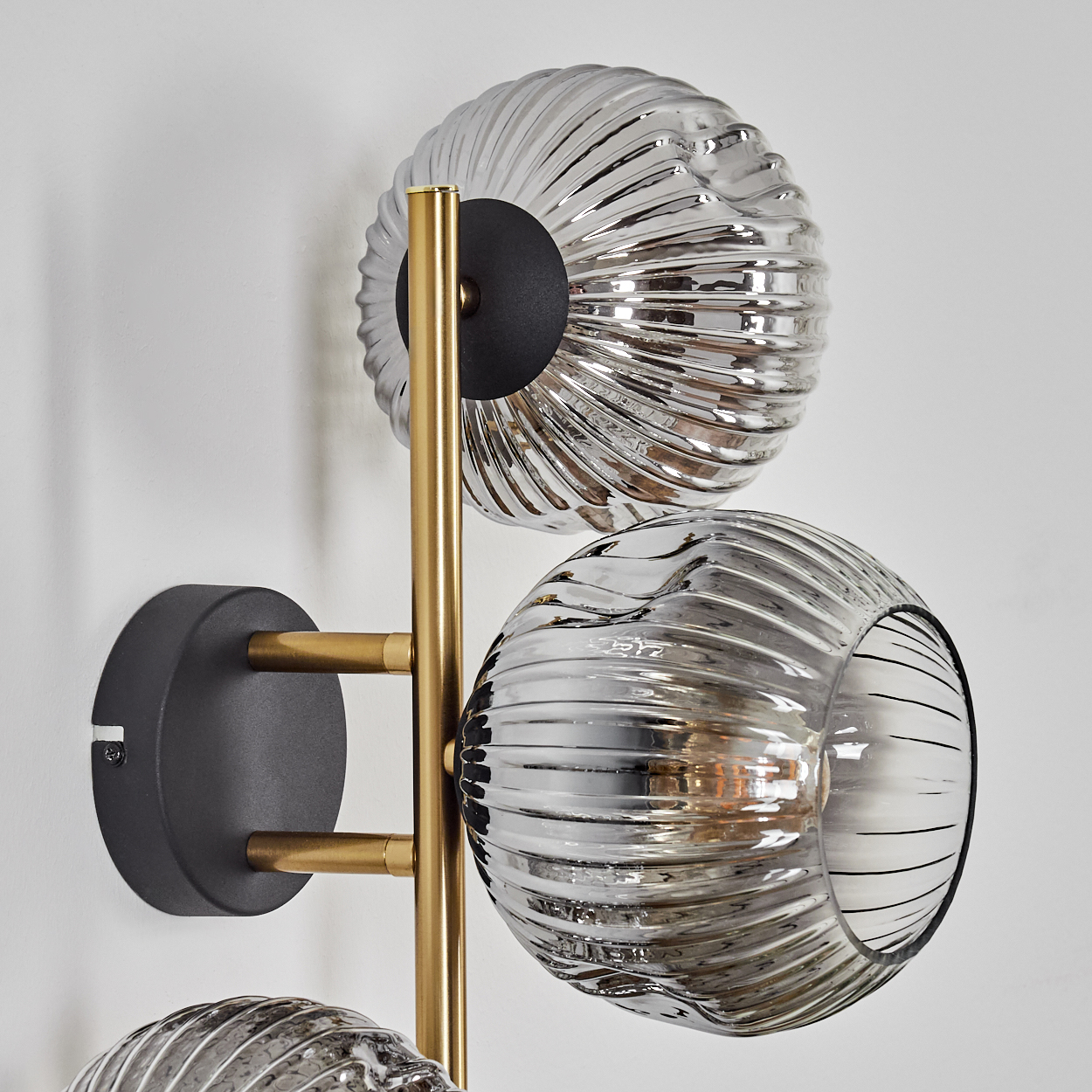 Eine moderne Wandlampe mit drei silbernen Glaskugeln, goldenen Metallarmen und einem schwarzen Sockel an einer weißen Wand.