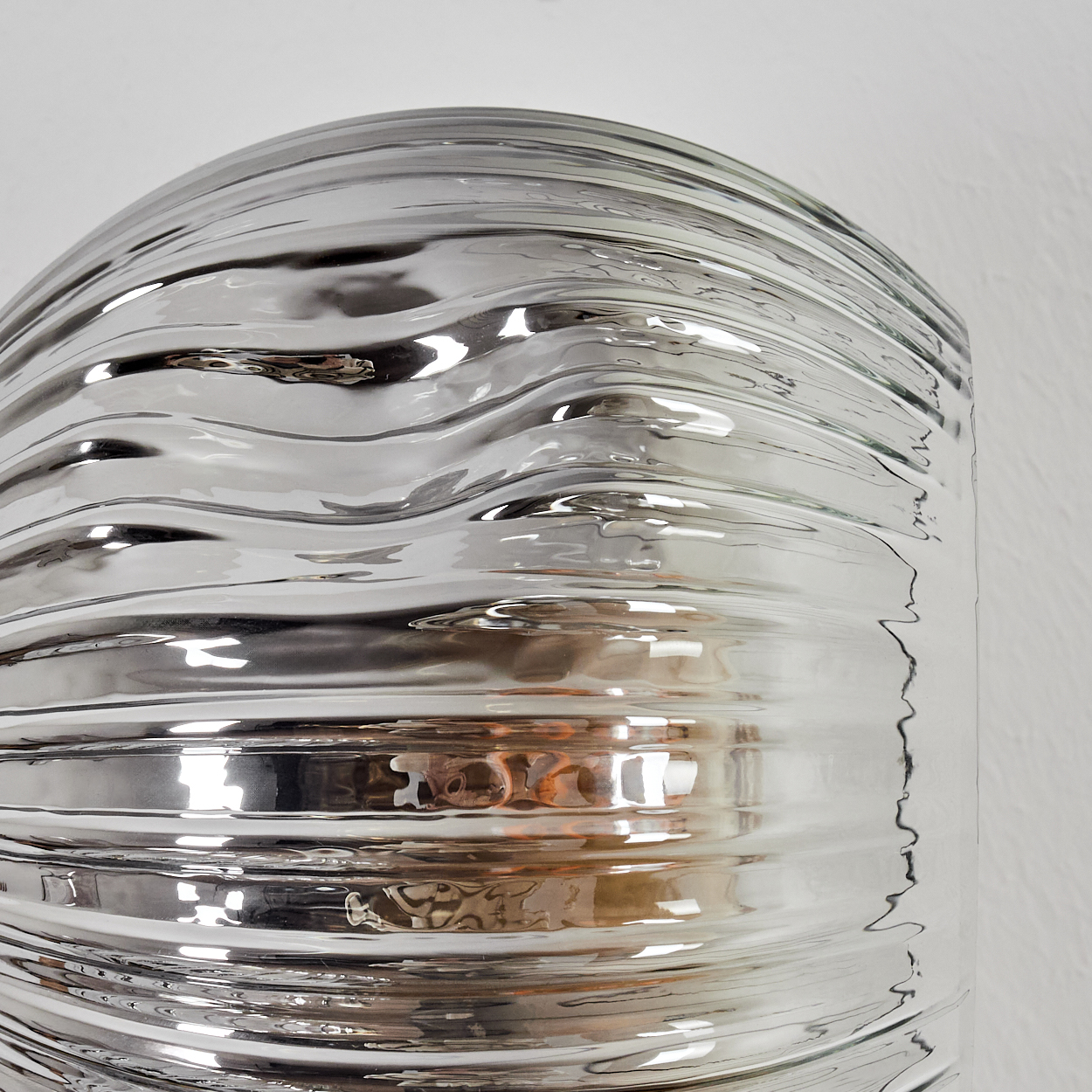 Nahaufnahme eines silbernen, gewellten Metallobjekts vor einer weißen Wand. Das Objekt reflektiert Licht mit orangefarbenen Reflexen.