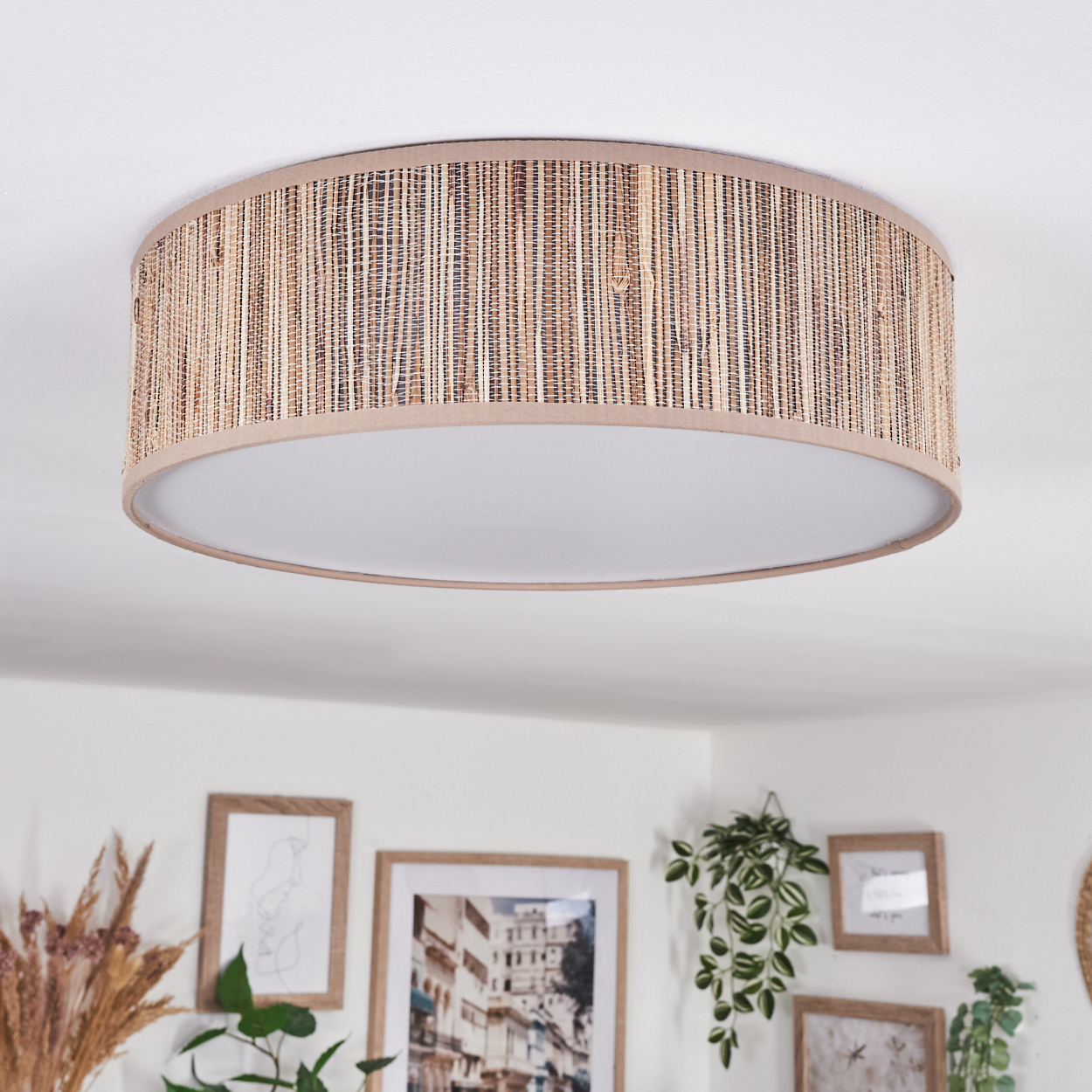 Eine runde Deckenlampe mit gewebtem Rattanschatten und weißem Innenraum. Im Hintergrund befinden sich mehrere gerahmte Bilder und Pflanzen.