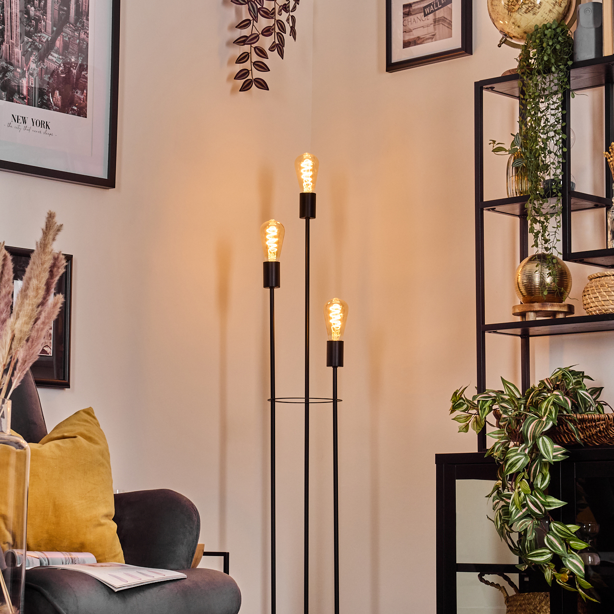 Ein modernes Wohnzimmer mit einer schwarzen Stehlampe mit drei Glühbirnen, einem dunkelgrauen Sessel und einem schwarzen Bücherregal mit Pflanzen.