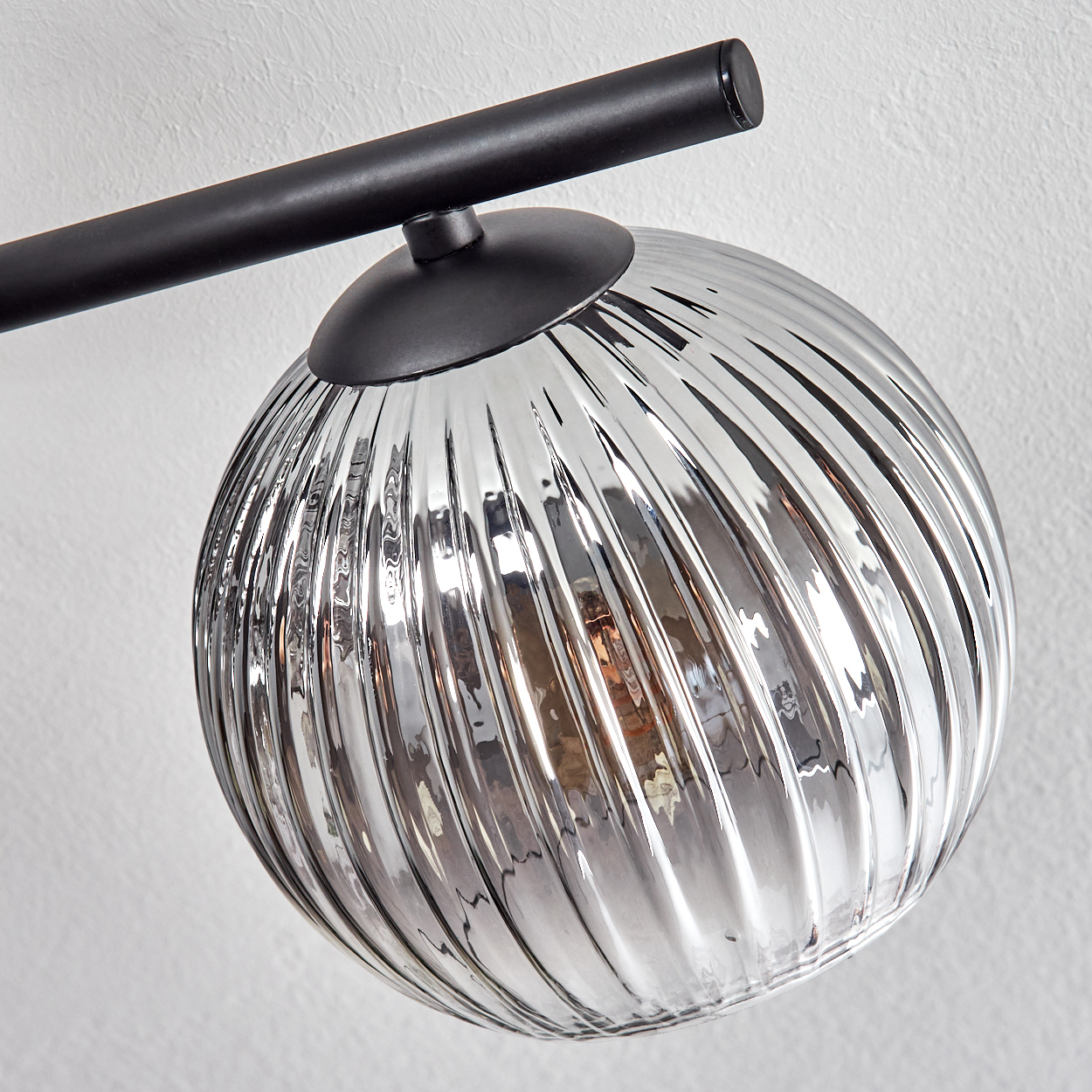 Nahaufnahme einer modernen Lampe. Sie hat einen schwarzen Arm, einen schwarzen Sockel und eine gerippte, silberne Glaskugel. Der Hintergrund ist eine weiße Wand.