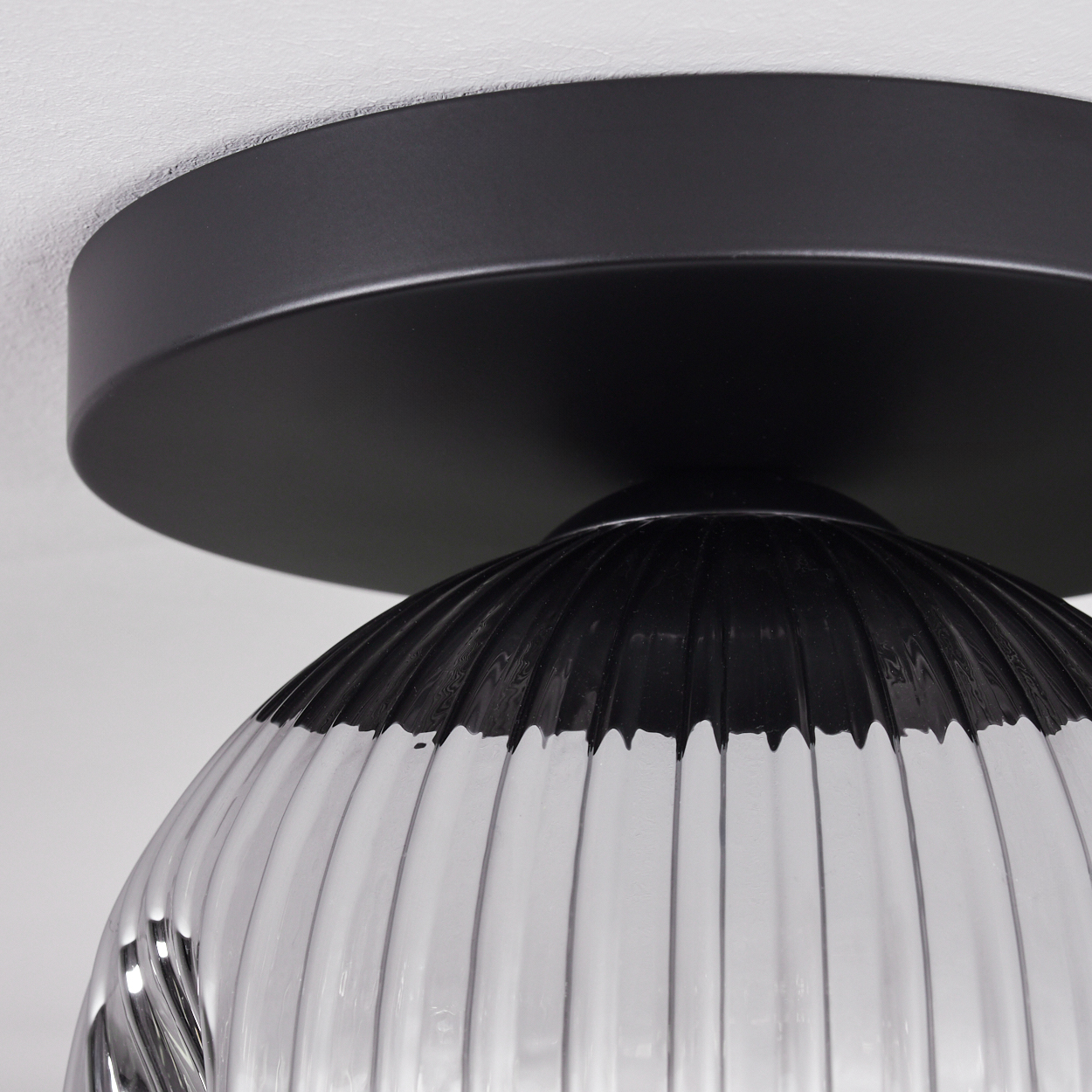 Nahaufnahme einer schwarzen Deckenlampe. Sie hat einen runden, schwarzen Sockel und einen gerippten, klaren Glasschirm. Der Hintergrund ist eine weiße Decke.