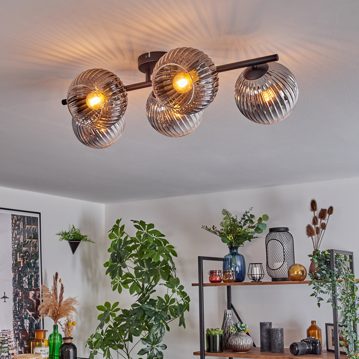Eine moderne Deckenleuchte mit Glaskugeln und ein Regal mit Pflanzen, Kerzen und Dekoration, das eine gemütliche Innenszene schafft.