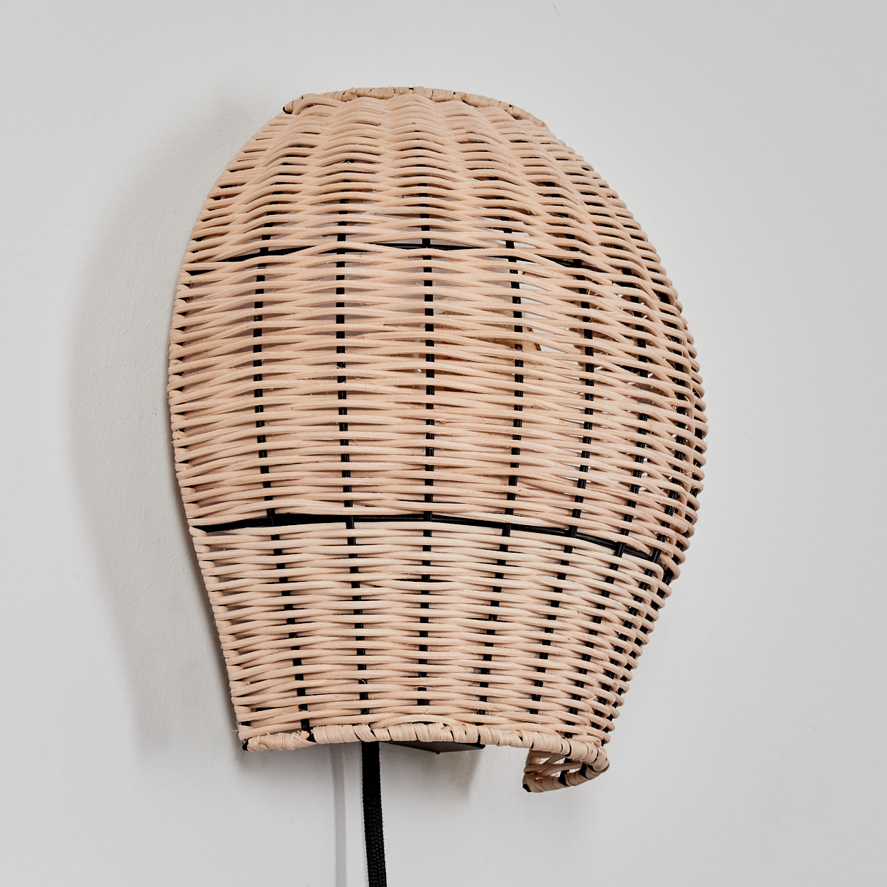 Wicker Lampe an weißer Wand. Die Lampe hat eine beige Farbe und ein schwarzes Kabel. Sie hat ein gewebtes Design.