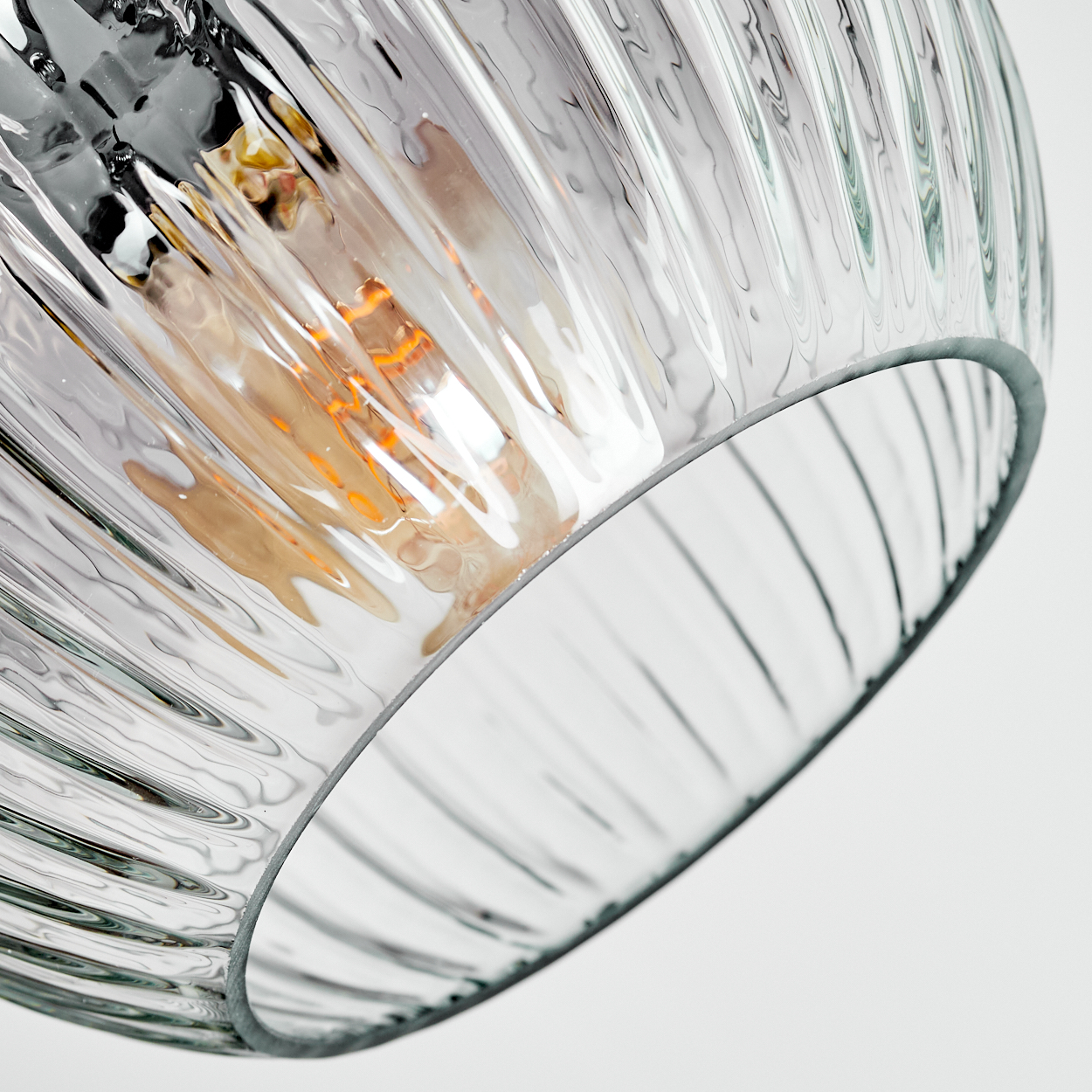 Nahaufnahme eines Lampenschirms aus klarem Glas mit vertikalen Rillen, der eine warm getönte Glühbirne beherbergt. Der Hintergrund ist schlicht weiß.