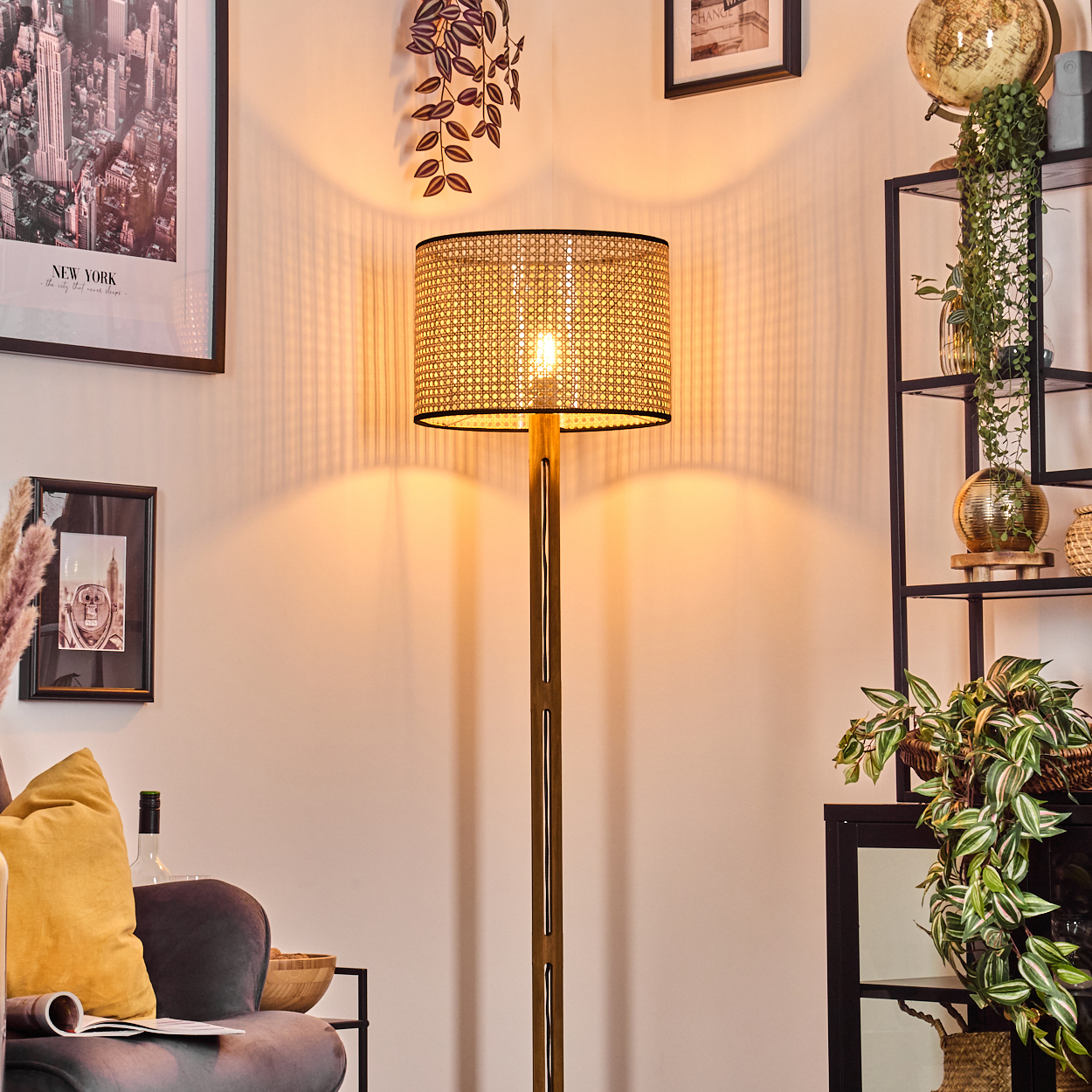 Eine beleuchtete Stehlampe mit gewebtem Schirm steht in einem Raum. Kunstwerke und Pflanzen schmücken den Raum, links ist eine Couch zu sehen.