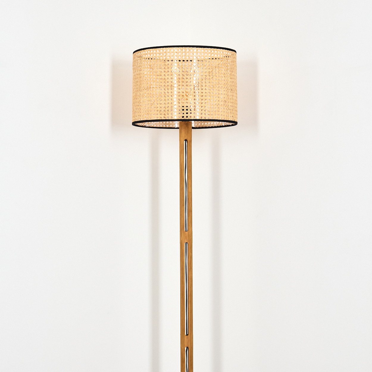 Eine Stehlampe mit gewebtem Lampenschirm und Holzstange steht vor einer weißen Wand. Der Schirm hat eine schwarze Umrandung.