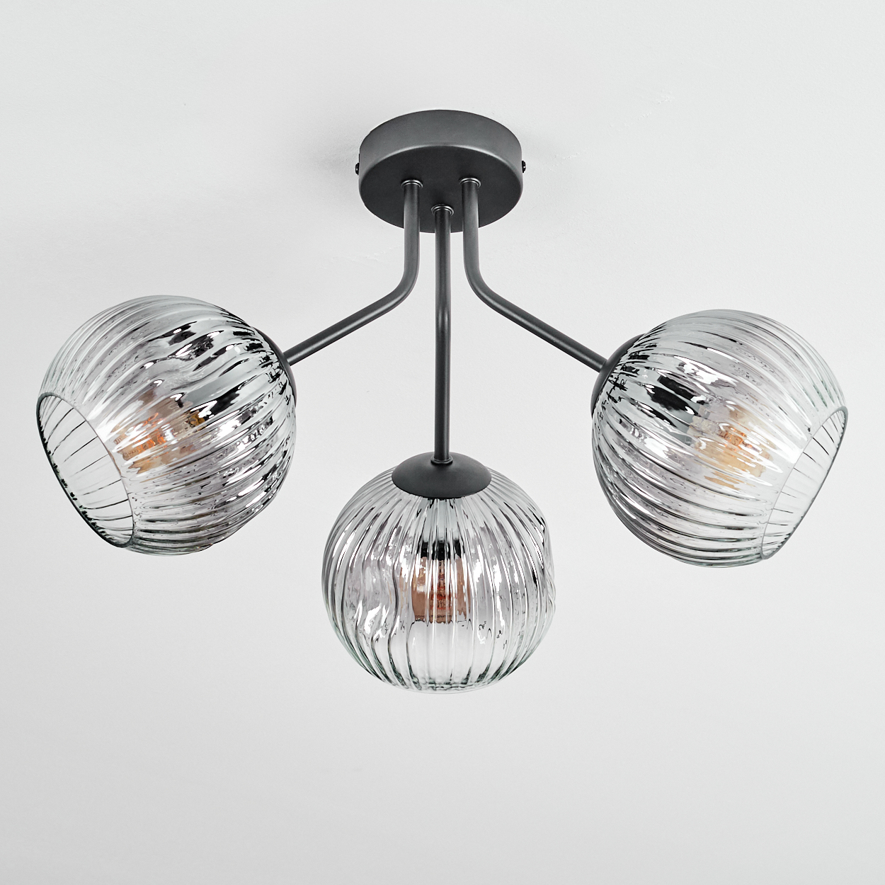 Eine moderne Deckenlampe mit drei kugelförmigen, gerippten Glasschirmen. Die Halterung ist dunkelgrau, vor weißem Hintergrund.