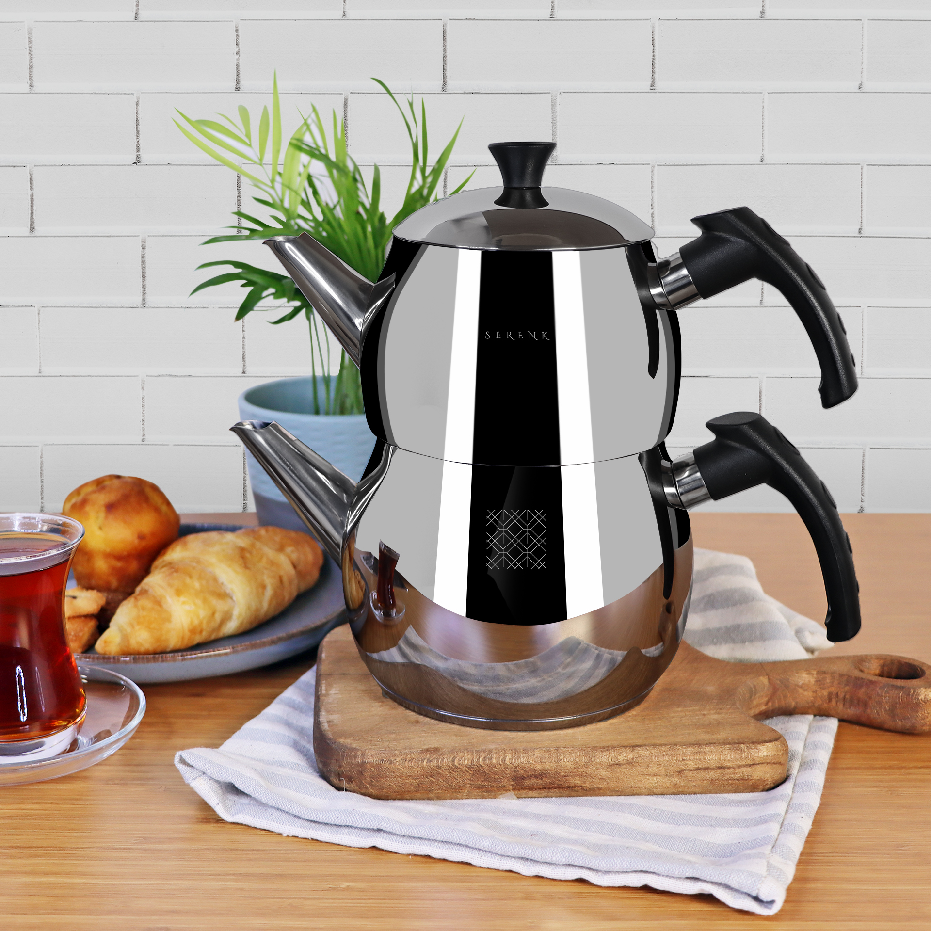 Una tetera cromada con asas negras, junto a una taza de té, pasteles y una planta. Sobre una mesa de madera.