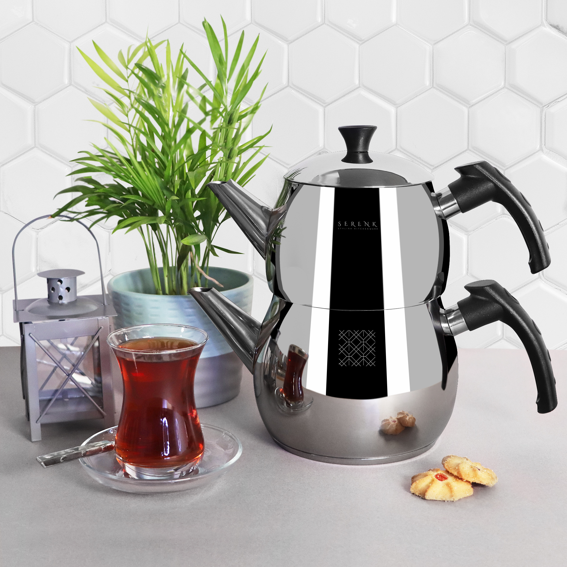 Un juego de té cromado con un vaso de té, una planta, una linterna y galletas sobre una superficie gris.