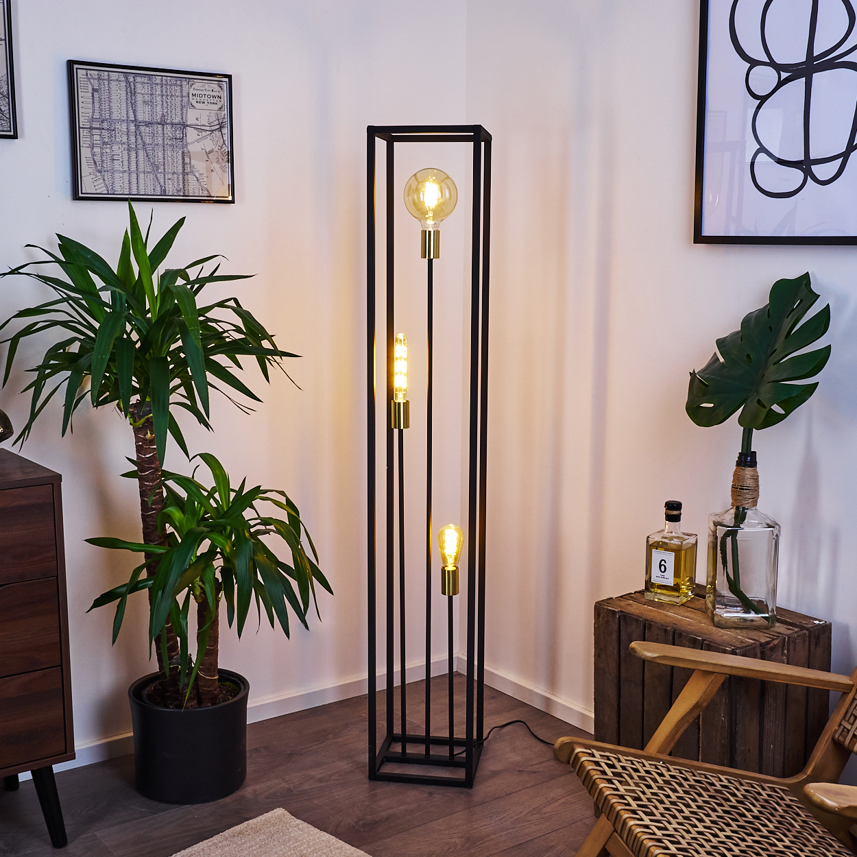 Ein modernes Zimmer mit einer schwarzen Metall-Stehlampe mit drei Glühbirnen. Eine Topfpflanze, eine Holzkiste und ein gerahmtes Kunstwerk sind ebenfalls vorhanden.